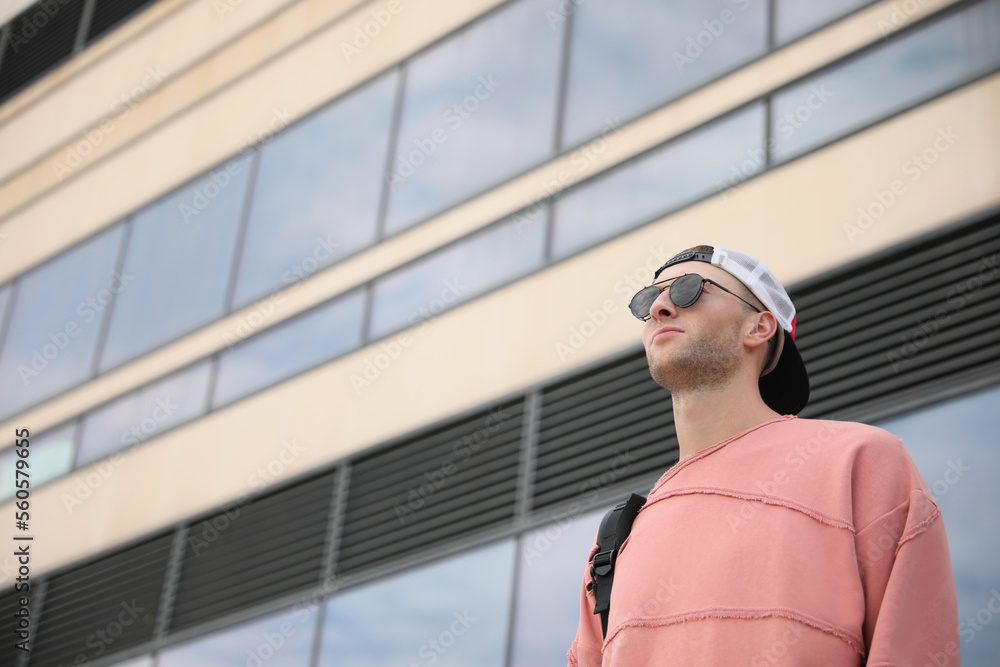 Handsome young man with stylish sunglasses and backpack near building outdoors, low angle view. Space for text Handsome young man with stylish sunglasses and backpack near building outdoors, low angle view. Space for text