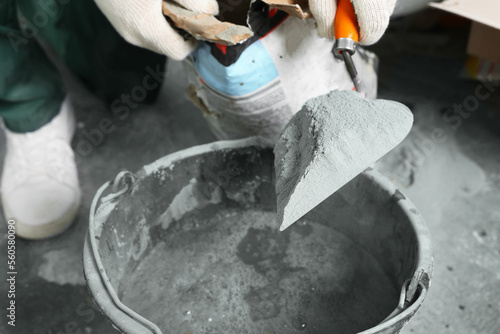 Worker with cement powder and trowel mixing concrete in bucket indoors, closeup