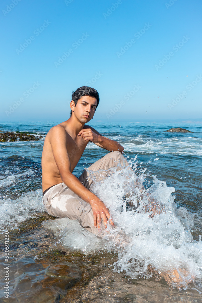 A dark-haired boy sits shirtless on the shore of the rocky beach with a wave crashing on his leg. Impressive water particles.