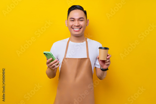 Smiling young Asian man barista barman employee wearing brown apron work in coffee shop holding mobile phoe and paper cup of coffee or tea isolated on yellow background. Small business startup photo