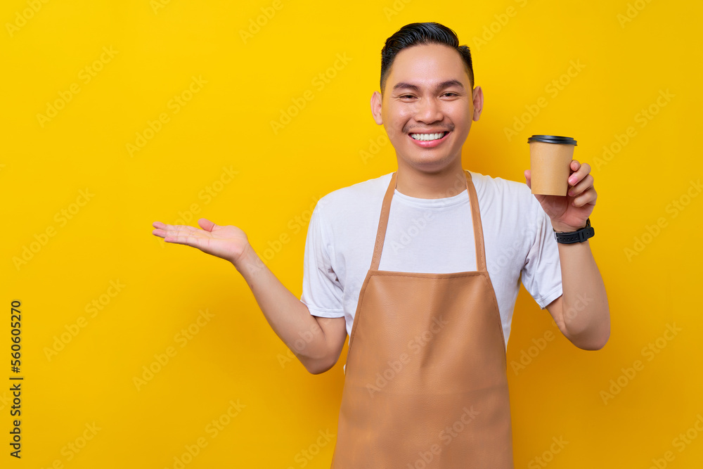 Excited young Asian man barista barman employee wearing brown apron work in coffee shop holding paper cup of coffee or tea and showing copy space on hand isolated on yellow background. Small business Excited young Asian man barista barman employee wearing brown apron work in coffee shop holding paper cup of coffee or tea and showing copy space on hand isolated on yellow background. Small business