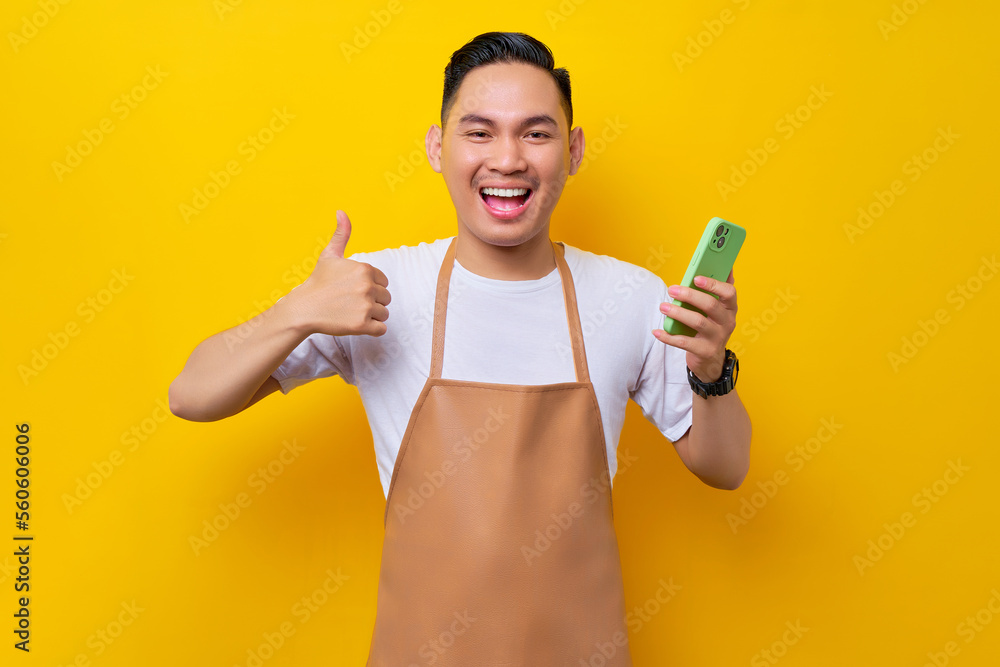 excited young Asian man barista barman employee wearing brown apron work in coffee shop holding mobile phone and showing thumb up sign isolated on yellow background. Small business startup concept excited young Asian man barista barman employee wearing brown apron work in coffee shop holding mobile phone and showing thumb up sign isolated on yellow background. Small business startup concept