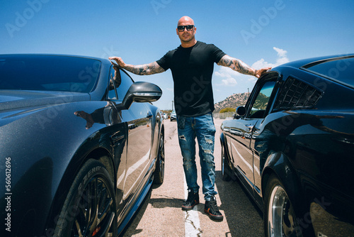 Man with shaved head standing in between two sports cars against a blue sky © F Armstrong Photo