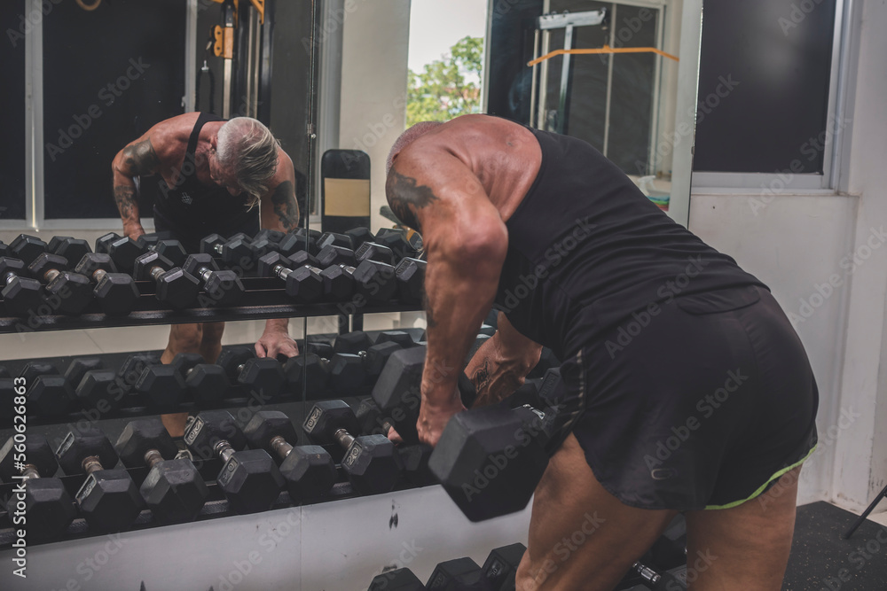 Man Straining As He Rows A muscular tanned older German guy does bent over one arm dumbbell rows ...