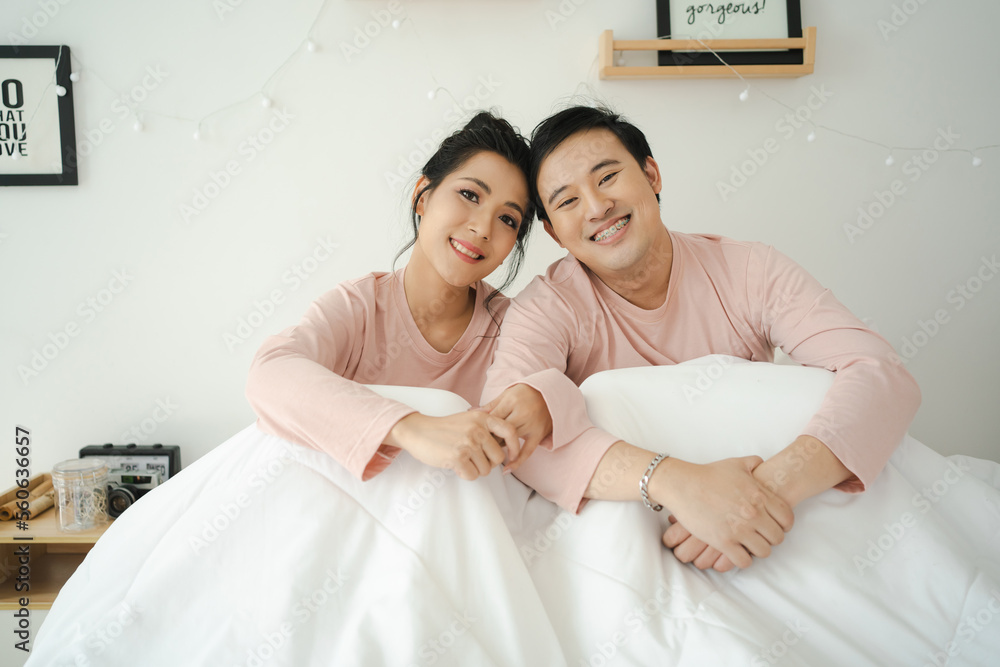 Fototapeta premium Happy and romantic Asian couple's portrait in bedroom with natural light from window, concept of relationship between husband and wife and being a family.