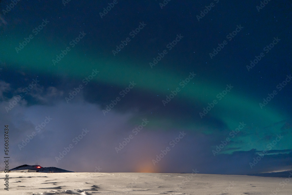 Aurora borealis with full moon lit clouds, glow of houses, construction crane and totally snowy ground with bluish starry sky in Iceland.