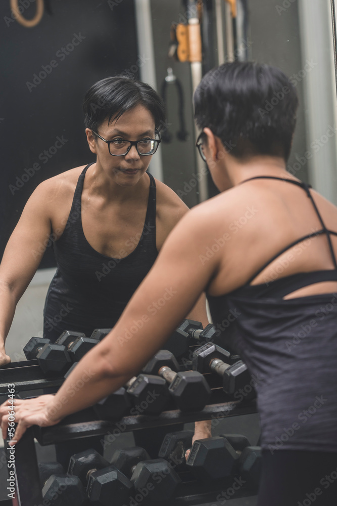 A fit middle aged asian woman with short hair getting into the zone before a workout. Picking up a dumbbell from the rack. Looking at her reflection in the gym.