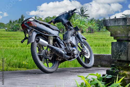 Old black motorbike on pavement with palm trees in background and blue skies with white clouds