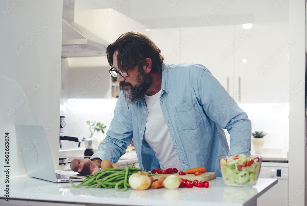 One man at home preparing fresh healthy salad using online tutorial. Modern people cooking vegetables looking for help on laptop and internet connection. Modern adult using computer to find recipes One man at home preparing fresh healthy salad using online tutorial. Modern people cooking vegetables looking for help on laptop and internet connection. Modern adult using computer to find recipes