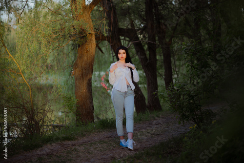 Sexy sensual brunette walks in a summer forest at sunset in a white blouse and jeans