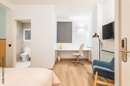 View of bedroom with large bed  work area with white wooden table and comfortable leather office chair. Small private bathroom with open door. To side is comfortable armchair with floor lamp.