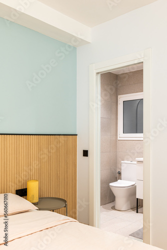 Open door to toilet room from cozy bedroom. Through doorway you can see white toilet bowl, tiled walls and floors, sink edge on black-legged vanity, and frosted plastic window.