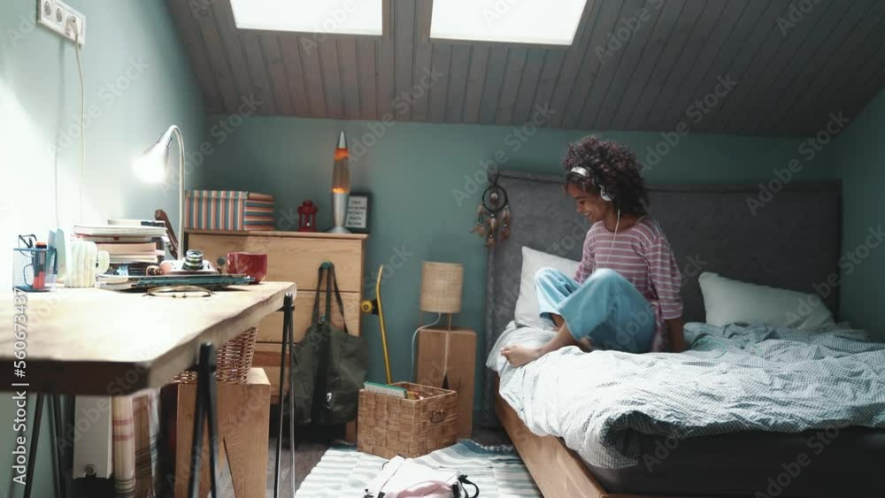 Handsome curly haired young African girl typing on mobile in headphones after walking on the bed at home Handsome curly haired young African girl typing on mobile in headphones after walking on the bed at home
