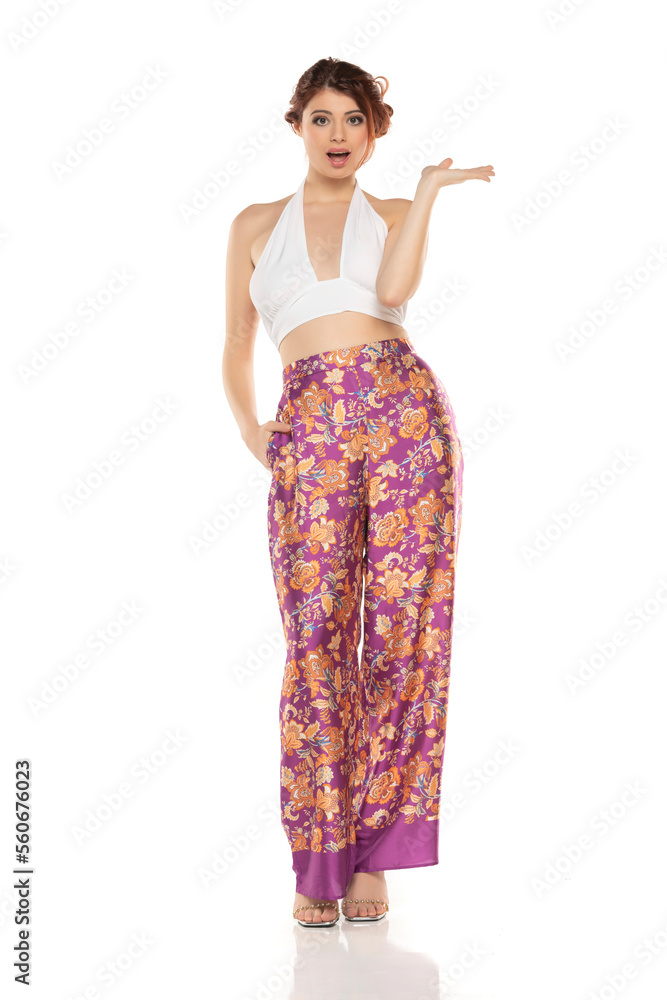 Smiling Redhead Woman In Floral Pants And Sleevles Shirt Presenting Something On her Empty Palm On A White Background