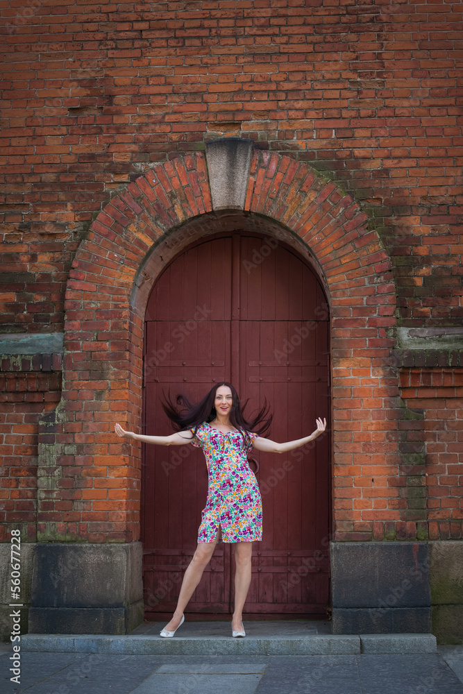 A beautiful slender smiling woman with long dark hair, in a light summer dress with a floral pattern, stands at the door in the form of an arch of an old red brick building.