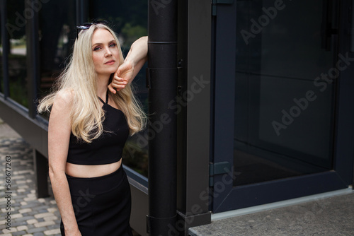 A stylish slender girl with blond long hair and a pleasant look stands against the backdrop of a modern building. Dressed in a short black sexy skirt and top. Sunglasses are on the head.