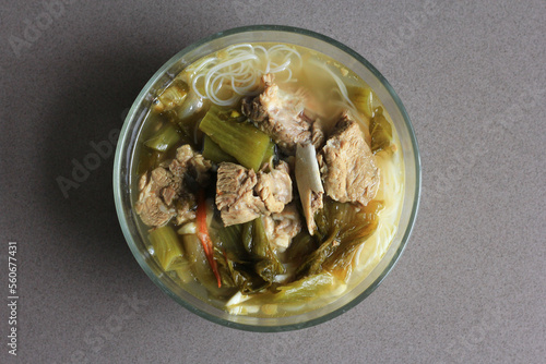 sup bakut or baikut babi sayur asin, is popular Chinese Indonesian pork rib soup with pickled cabbage or mustard greens and vermicelli in clear bowl, on black background, top view.