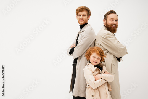 joyful bearded man in trench coats standing back to back with crossed arms near redhead boy isolated on grey.
