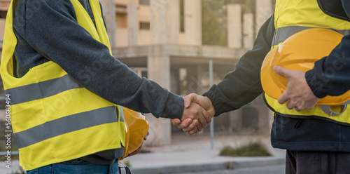 Bild auf Leinwand Dos operarios de construcción, estrechándose la mano y de fondo un edificio en obra