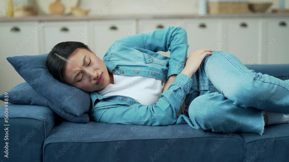 Sick young asian woman feeling stomach ache while lying on sofa in living room at home. Tired female on the couch has an upset stomach, acute gastritis or menstruation, a feeling of spasms, heartburn Sick young asian woman feeling stomach ache while lying on sofa in living room at home. Tired female on the couch has an upset stomach, acute gastritis or menstruation, a feeling of spasms, heartburn