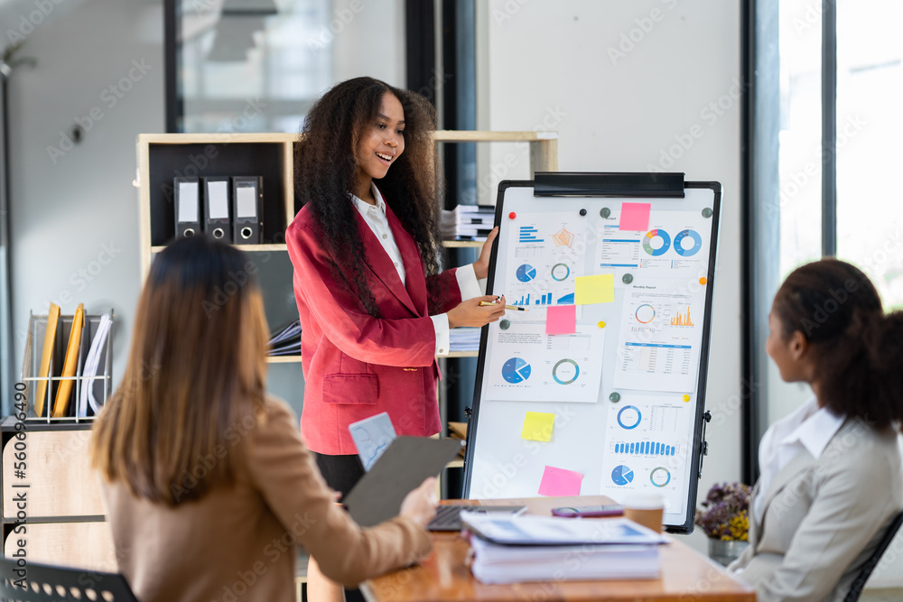 A group of young American businesswomen pointed to a whiteboard to provide information and a portfolio chart for colleagues to comment on and smile, proud of their creations. A group of young American businesswomen pointed to a whiteboard to provide information and a portfolio chart for colleagues to comment on and smile, proud of their creations.