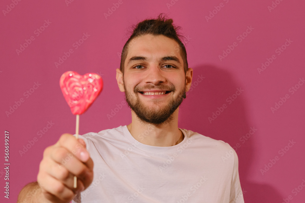 Young Caucasian man with beard shows sweet red lollipop in shape of heart and smiles. Isolated on pink background. Full-face portrait. Concept of Valentine's Day. Young Caucasian man with beard shows sweet red lollipop in shape of heart and smiles. Isolated on pink background. Full-face portrait. Concept of Valentine's Day.