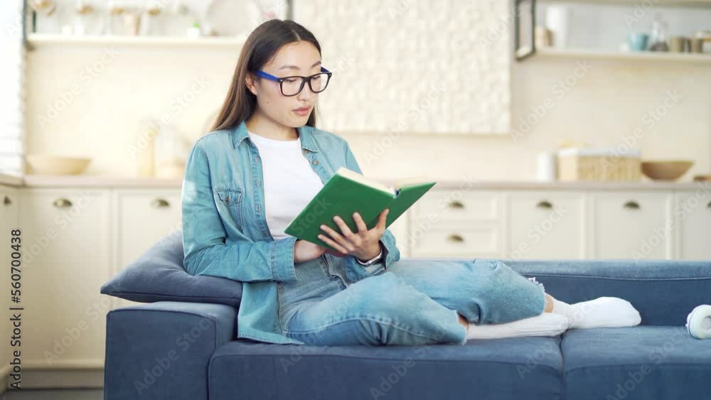 Young Asian woman wearing glasses reading a book while sitting or lying on sofa in living room. A female in casual clothes is holding a study guide while relaxing on the couch in the kitchen at home Young Asian woman wearing glasses reading a book while sitting or lying on sofa in living room. A female in casual clothes is holding a study guide while relaxing on the couch in the kitchen at home