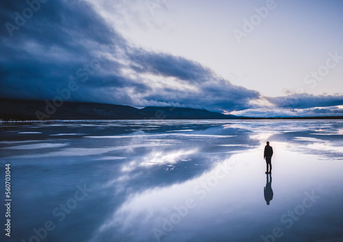 Lone figure stands on frozen lake with reflection and storm clouds