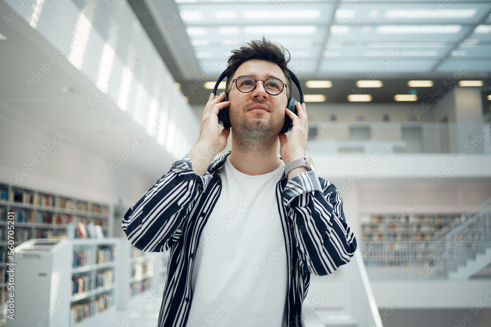 Fototapeta premium Student, library and man with music headphones in university, college or school. Thinking, education scholarship and face of young male streaming educational podcast, radio or song, audio or album.