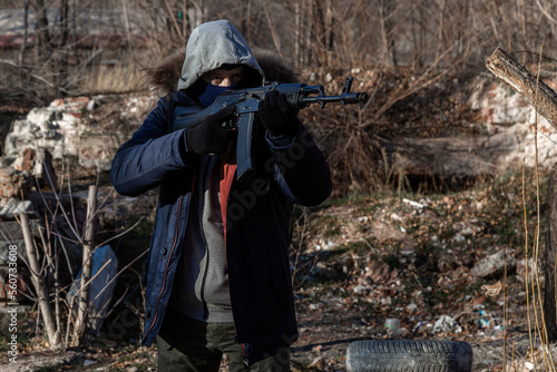 A grown man wearing a mask, wearing a jacket with a hood, wearing military pants, wearing gloves aims at a weapon