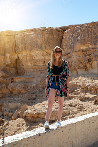 The model walks along the observation deck against the background of rocks in sunny weather. The girl is dressed like a tourist: sunglasses, blue shorts, a T-shirt and a Hawaiian shirt.