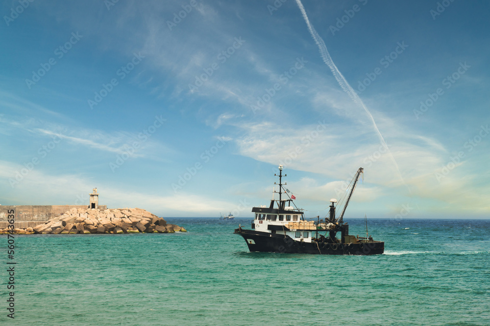 View of a fishing vessel returning from the sea to land and a view of the small harbor, lighthouse and gorgeous dramatic sky. 