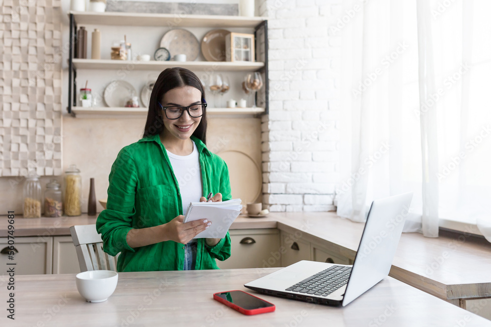A young female journalist, writer, blogger works remotely from home using a laptop online. Sitting in the kitchen at the table, making notes in a notebook. A young female journalist, writer, blogger works remotely from home using a laptop online. Sitting in the kitchen at the table, making notes in a notebook.
