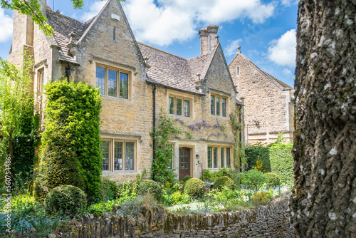 A honey coloured stone cottage with 2 gables overlooking the front garden and river Windrush in the Cotswold village of Bourton on the Water in Gloustershire England