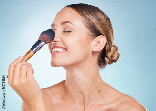 Woman, face and smile with makeup brush for skincare beauty, dermatology wellness and cosmetics treatment in blue background. Model happiness, facial care and luxury product application in studio