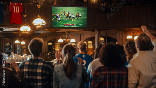 Group of American Football Fans Watching a Live Match Broadcast in a Sports Pub on TV. People Cheering, Supporting Their Team. Crowd Goes Ecstatic When Team Scores a Goal and Wins the Championship. © Gorodenkoff