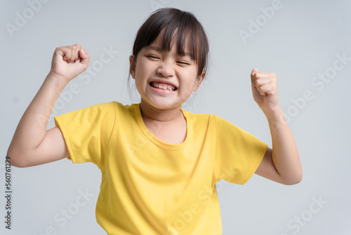 Cute Asian girl, 6 to 7 years old, black hair, white sleeveless shirt. She clenched her fists and raised both of her arms.To show the muscles in the arms and their strength with a happy smiling face.