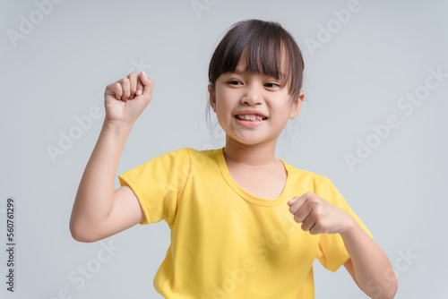 Cute Asian girl, 6 to 7 years old, black hair, white sleeveless shirt. She clenched her fists and raised both of her arms.To show the muscles in the arms and their strength with a happy smiling face.