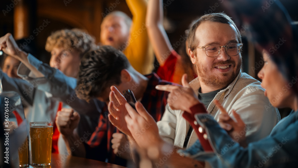 Obraz premium Portrait of an Excited Young Man Sitting at Bar Counter, Drinking Beer and Using a Smartphone, Anxious About a Sports Bet on His Favorite Soccer Team. Joyful Emotions When Football Team Scores a Goal.