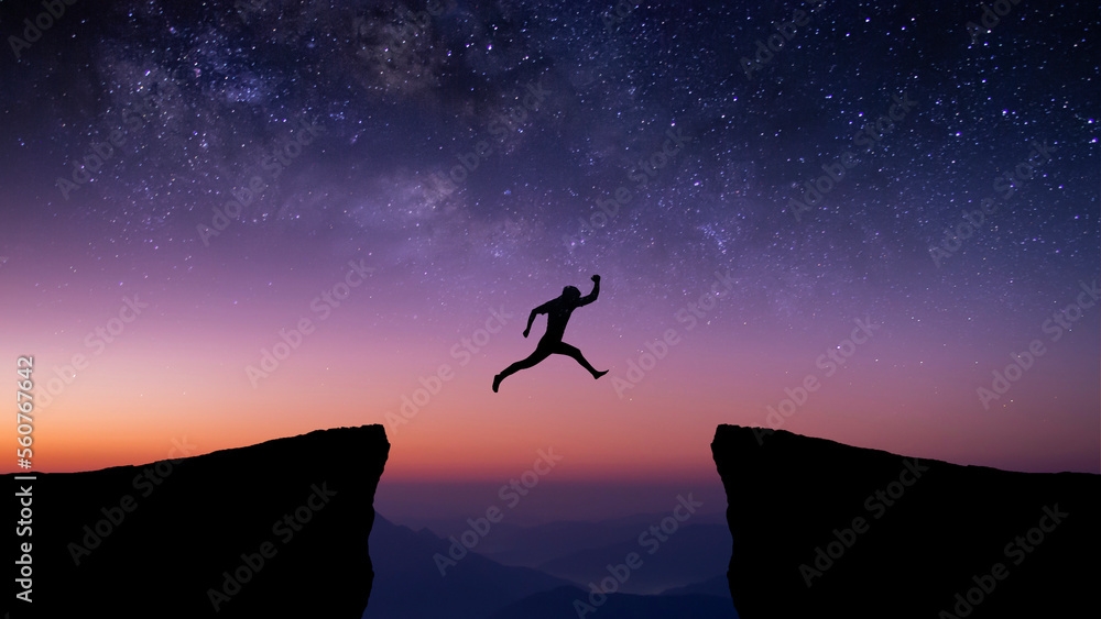 Silhouette of young traveler and backpacker jumping on cliff at night with the star and milky way over the sky. He enjoyed traveling and was successful when he reached the summit.