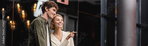 joyful blonde woman looking away and pointing with finger near boyfriend in hotel hall, banner.