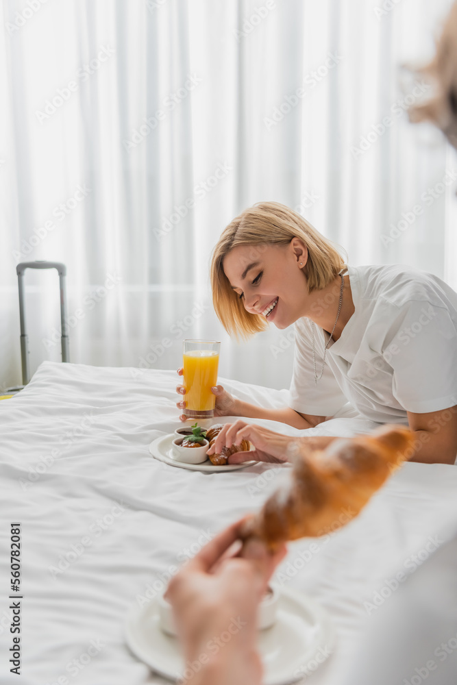 cheerful blonde woman having breakfast with orange juice and croissant on hotel bed near blurred boyfriend. cheerful blonde woman having breakfast with orange juice and croissant on hotel bed near blurred boyfriend.