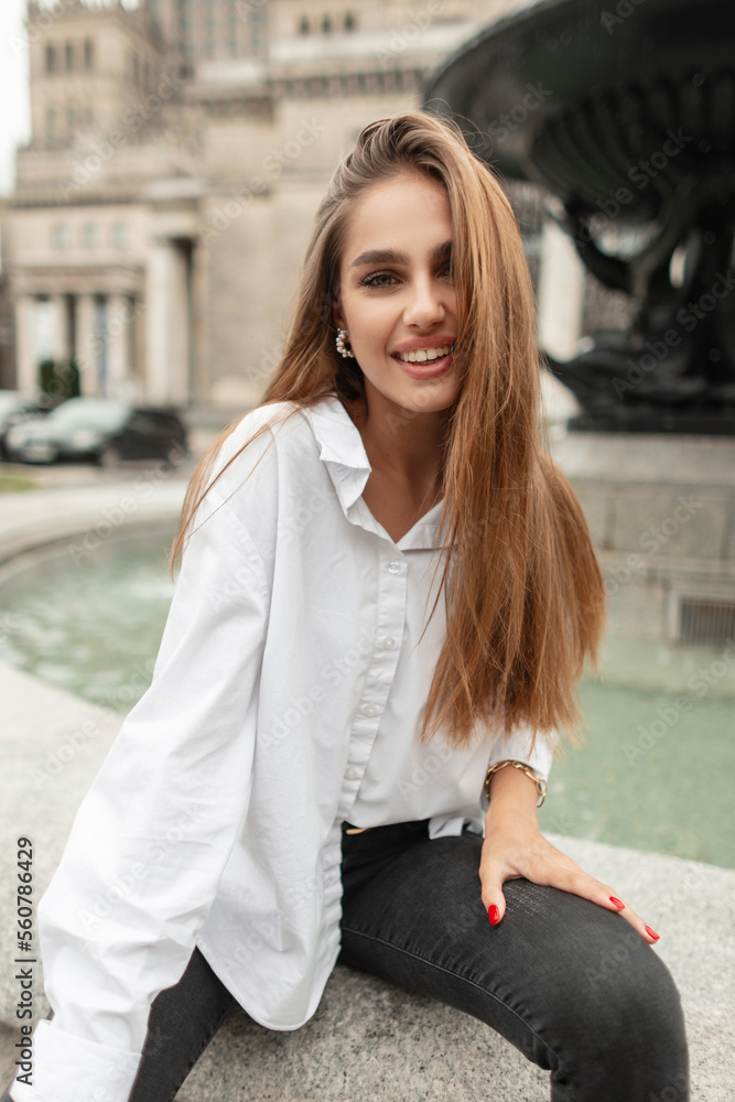 Happy beautiful young girl with a smile in fashionable casual clothes with a white shirt and black jeans sits near a vintage fountain in a European town