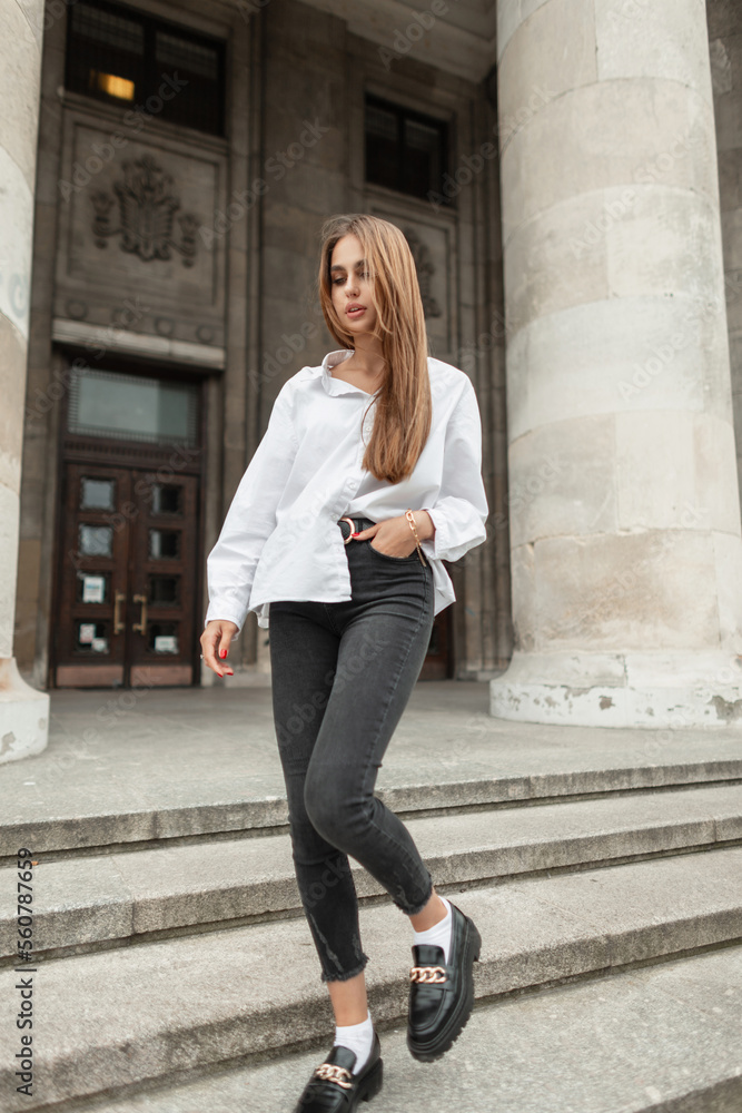 Fashionable beautiful business woman in a stylish clothes with a white shirt, black jeans, shoes walking near a vintage building in the city