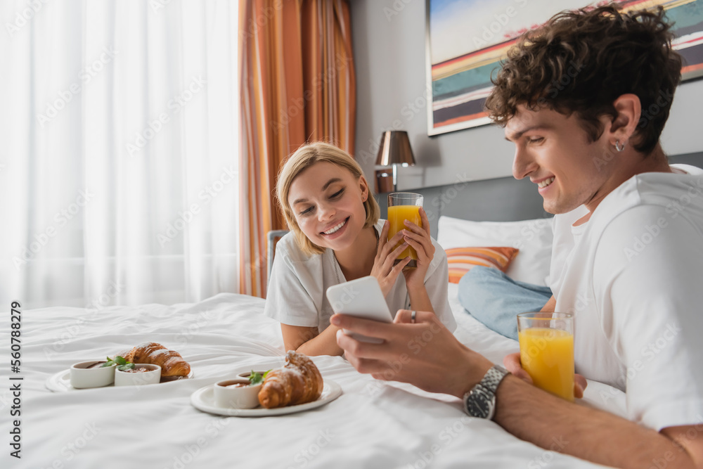 young man showing mobile phone to smiling girlfriend near croissants and orange juice on hotel bed.