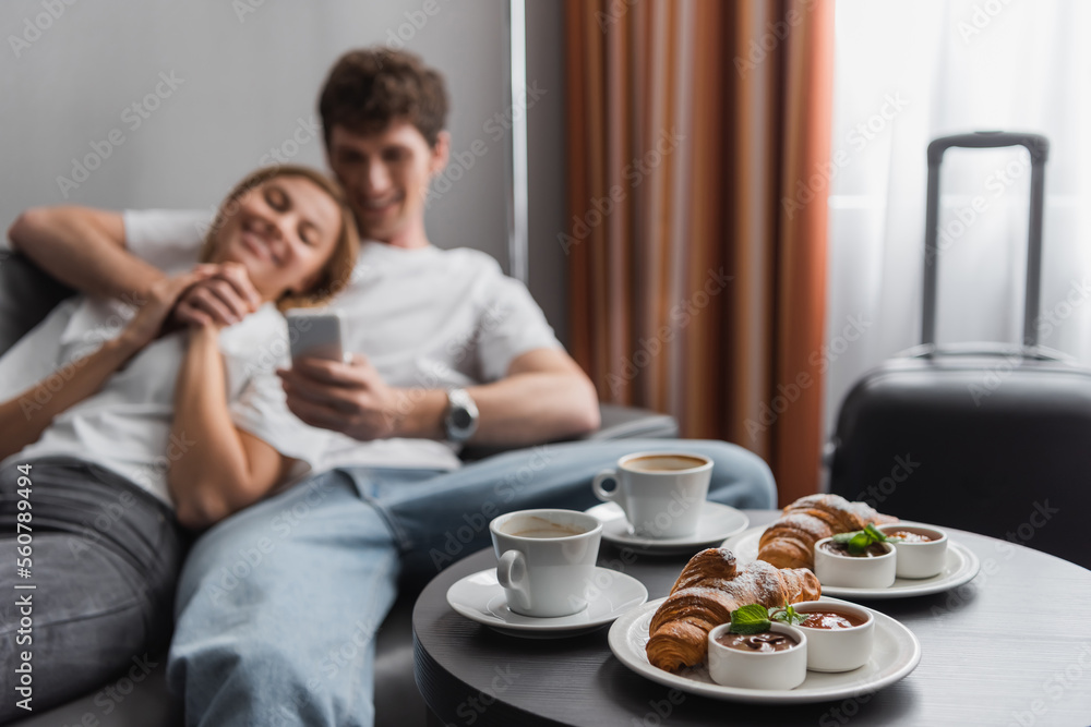 coffee cups and croissants with jam and chocolate paste near blurred couple with smartphone in hotel room. coffee cups and croissants with jam and chocolate paste near blurred couple with smartphone in hotel room.