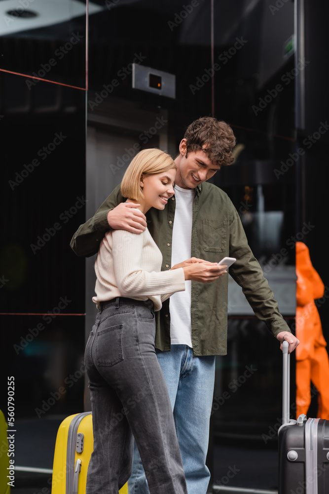 Obraz premium smiling woman showing mobile phone to boyfriend embracing her near baggage in hotel lobby.