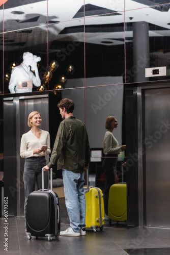 young blonde woman with smartphone smiling near boyfriend with suitcases in lobby of modern hotel.