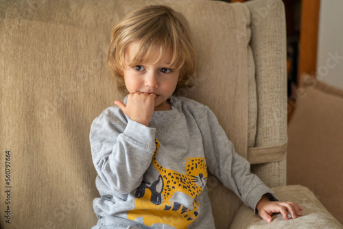 Fotografia Blond Preschool Boy Sitting On The Sofa At Home, Biting His Nails