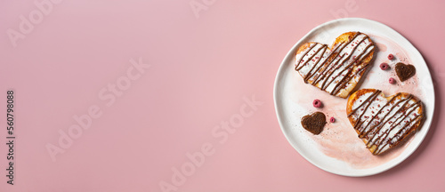 Valentine's Day wide banner with copy space. Heart-shaped French toast with cream, chocolate, and freeze-dried raspberry crumbles, on a ceramic plate against a light pink background.